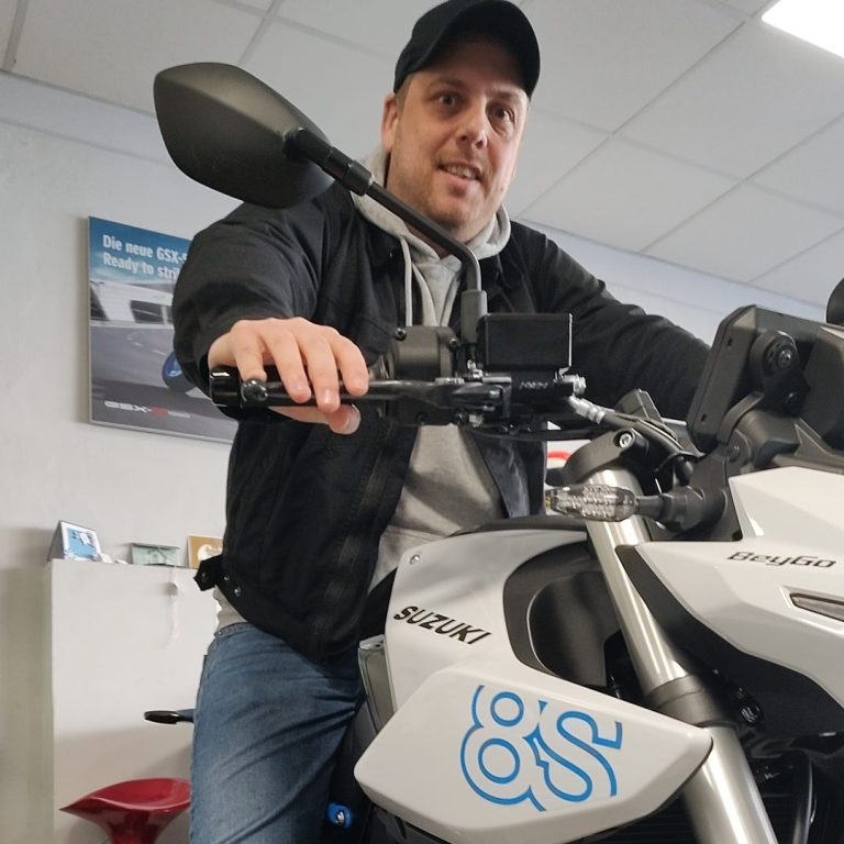 Mann sitzt auf einem Motorrad in einem Verkaufsraum, lächelt und trägt eine Kappe.
